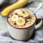 Warm banana mug cake topped with banana slices and chocolate chips served in a ceramic mug with a spoon.
