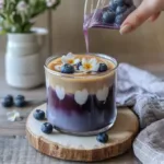 Iced blueberry latte layered with blueberry syrup, milk, and espresso, garnished with fresh blueberries and chamomile flowers on a wooden board