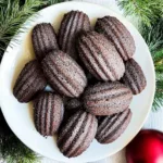 A plate of rich chocolate madeleines with a slightly crisp edge and soft, moist center, arranged neatly on a white plate, surrounded by festive pine branches and a red ornament.