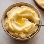 A bowl of creamy Honey Butter recipetopped with a drizzle of golden honey and a spoon resting inside, showcasing its smooth and fluffy texture.