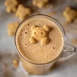 A warm Teddy Graham latte topped with a honey graham cookie floating on creamy froth, showing cozy cinnamon coffee vibes perfect nostalgic fall drink.