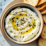 A bowl of creamy whipped ricotta dip drizzled with olive oil and sprinkled with herbs and black pepper, served with toasted bread slices.