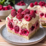 Moist white chocolate raspberry cake slice topped with fresh raspberries and creamy frosting on a plate.